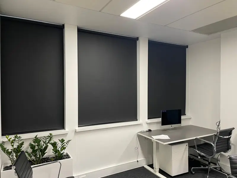 Office with dark blockout roller blinds fitted to a wall of windows