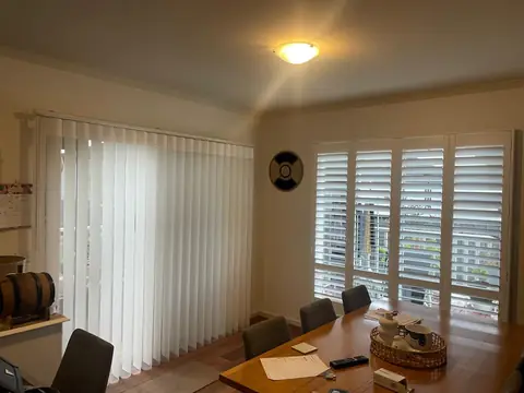 White vertical blinds across a dining room's sliding door, with plantation shutters on the window beside